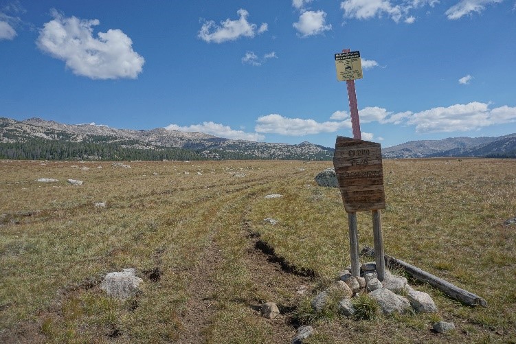 Cloud Peak Wilderness sign with a large flat grassy field with boulders and a forest and mountains in the background.